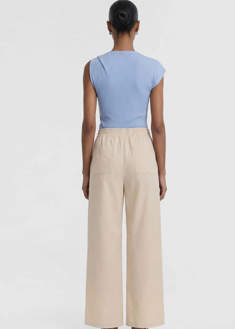 A model wearing a light blue knit top with a unique asymmetrical shoulder tie detail and beige drawstring trousers, shot against a plain light grey background.