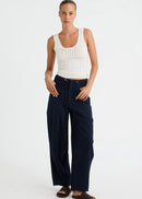 A woman modelling a white ribbed tank top and navy wide-leg trousers with dark brown slide sandals, standing against a light grey studio background.