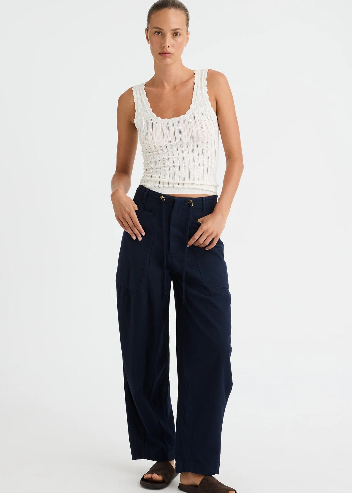 A woman modelling a white ribbed tank top and navy wide-leg trousers with dark brown slide sandals, standing against a light grey studio background.