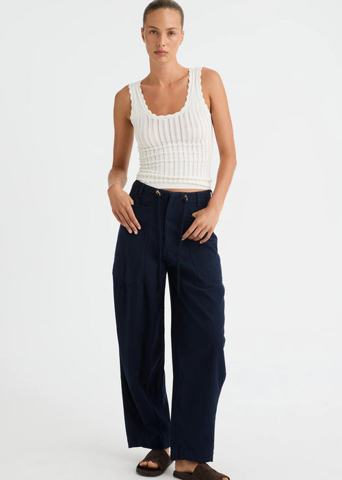 A woman modelling a white ribbed tank top and navy wide-leg trousers with dark brown slide sandals, standing against a light grey studio background.