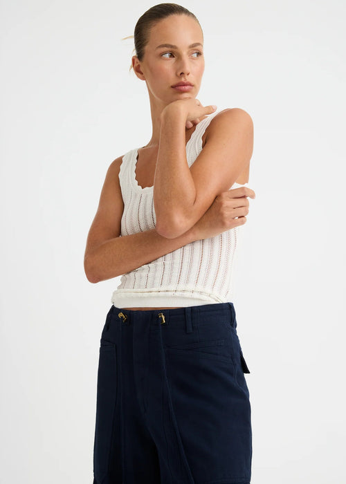 A woman modelling a white ribbed sleeveless top and navy blue trousers against a plain white studio background.