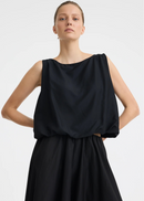 A woman modelling a sleeveless black dress with a draped, voluminous bodice and a flowing skirt against a plain light grey studio background.