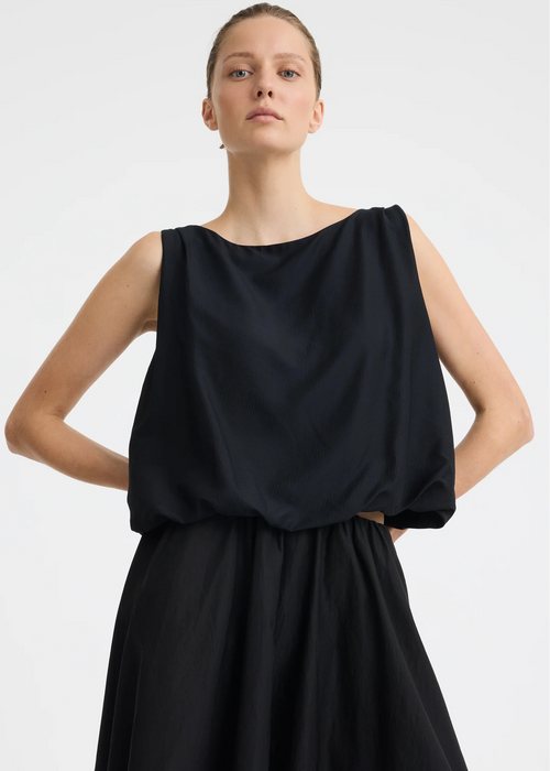 A woman modelling a sleeveless black dress with a draped, voluminous bodice and a flowing skirt against a plain light grey studio background.
