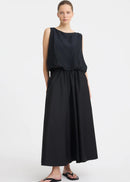 A woman modelling a sleeveless black dress with a draped, voluminous bodice and a flowing skirt against a plain light grey studio background.