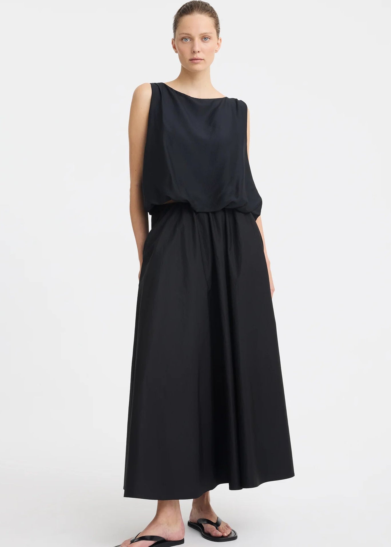 A woman modelling a sleeveless black dress with a draped, voluminous bodice and a flowing skirt against a plain light grey studio background.