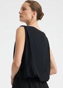 A woman modelling a sleeveless black dress with a draped, voluminous bodice and a flowing skirt against a plain light grey studio background.