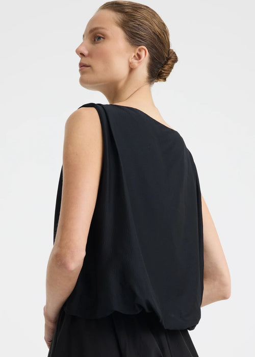 A woman modelling a sleeveless black dress with a draped, voluminous bodice and a flowing skirt against a plain light grey studio background.