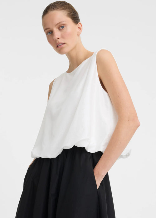 A woman modelling a sleeveless white top with a high neckline and black skirt against a light grey studio background.