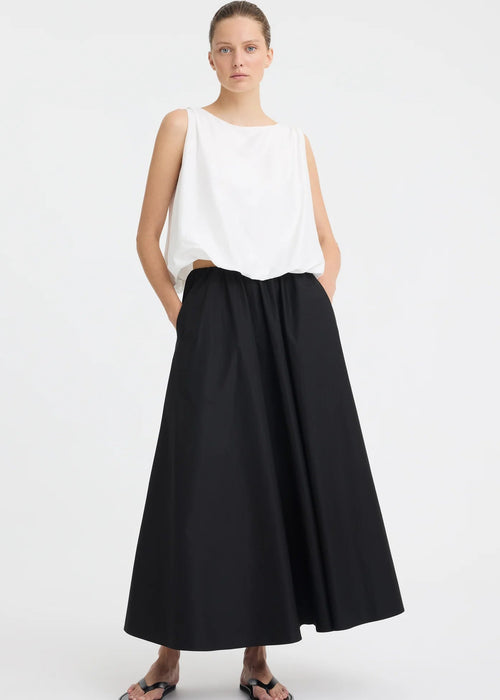 A woman modelling a sleeveless white top with a high neckline and black skirt against a light grey studio background.