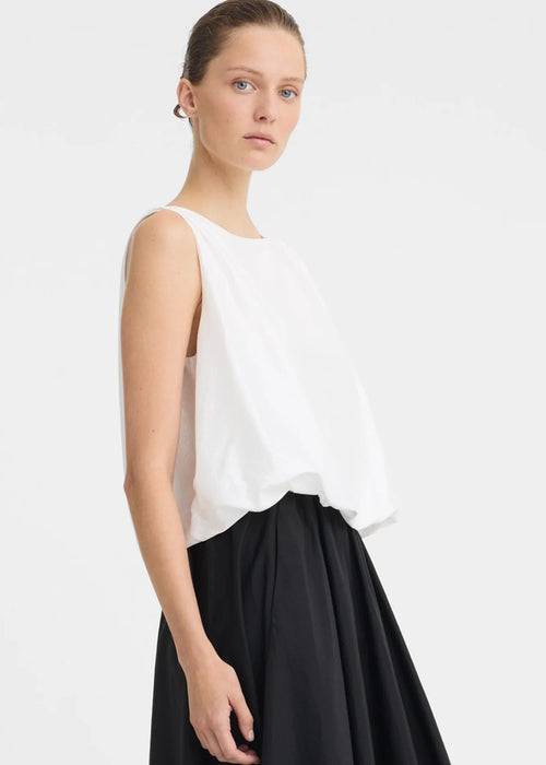 A woman modelling a sleeveless white top with a high neckline and black skirt against a light grey studio background.