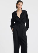 A woman modelling a black long-sleeve wrap-style blouse and matching wide-leg trousers, standing against a plain light grey studio background.