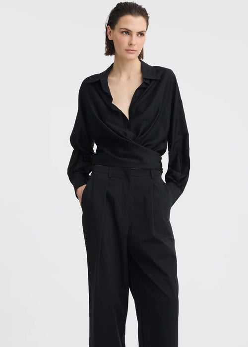 A woman modelling a black long-sleeve wrap-style blouse and matching wide-leg trousers, standing against a plain light grey studio background.