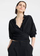 A woman modelling a black long-sleeve wrap-style blouse and matching wide-leg trousers, standing against a plain light grey studio background.