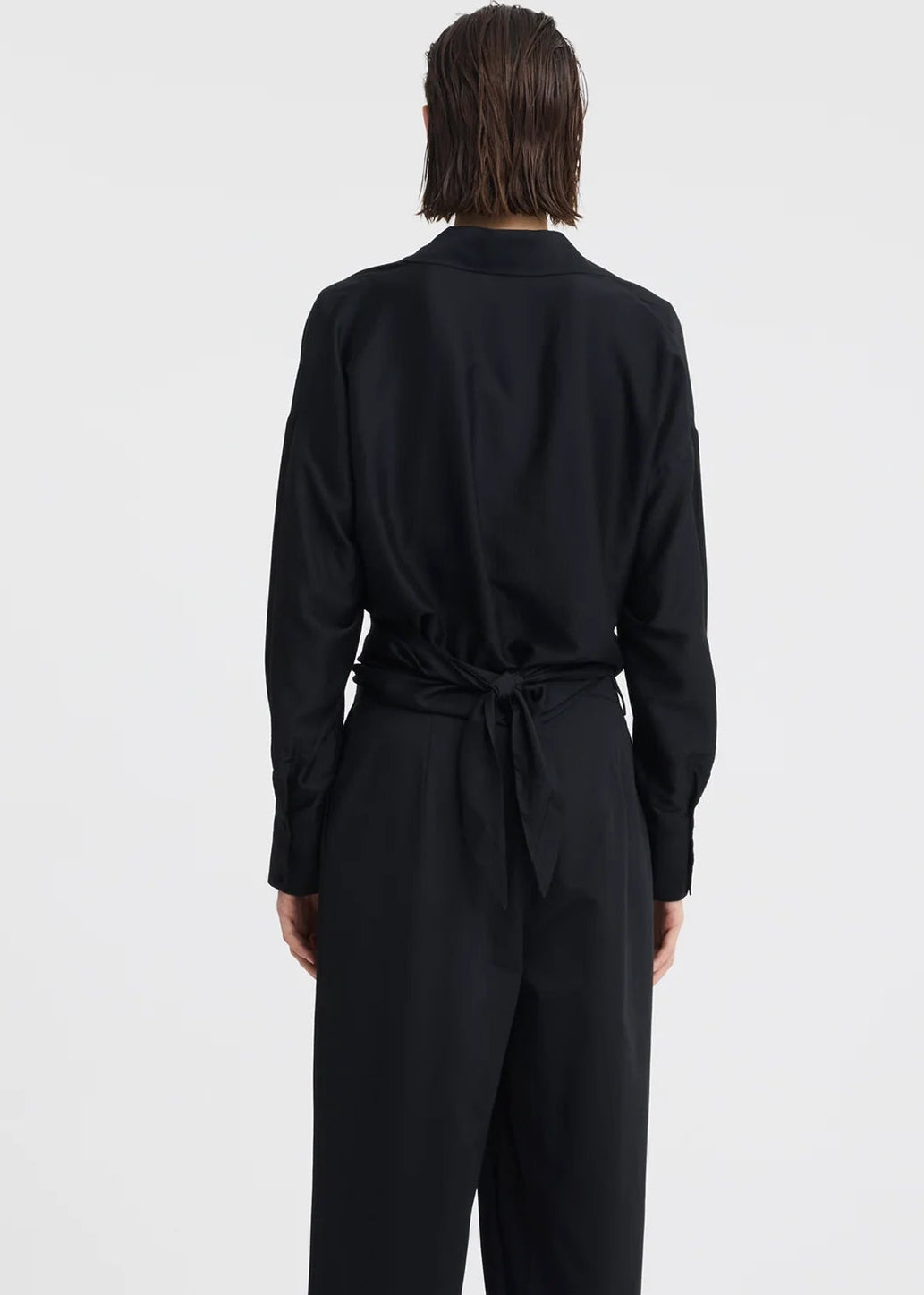 A woman modelling a black long-sleeve wrap-style blouse and matching wide-leg trousers, standing against a plain light grey studio background.