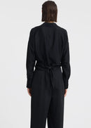 A woman modelling a black long-sleeve wrap-style blouse and matching wide-leg trousers, standing against a plain light grey studio background.