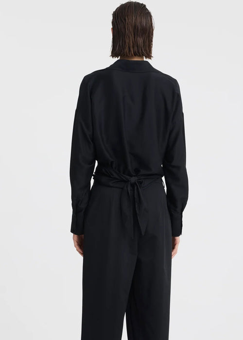 A woman modelling a black long-sleeve wrap-style blouse and matching wide-leg trousers, standing against a plain light grey studio background.