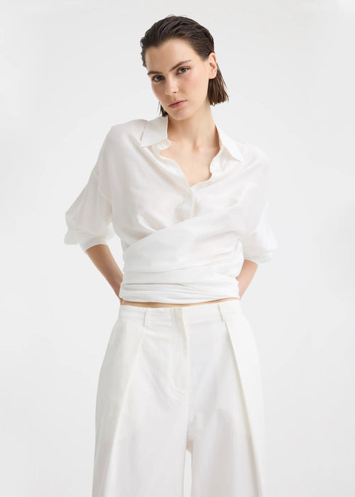 A model wearing a white long-sleeve button-up shirt and matching white wide-leg trousers, styled with black flip-flop sandals against a plain white studio background.