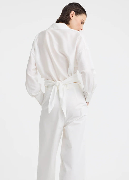 A model wearing a white long-sleeve button-up shirt and matching white wide-leg trousers, styled with black flip-flop sandals against a plain white studio background.