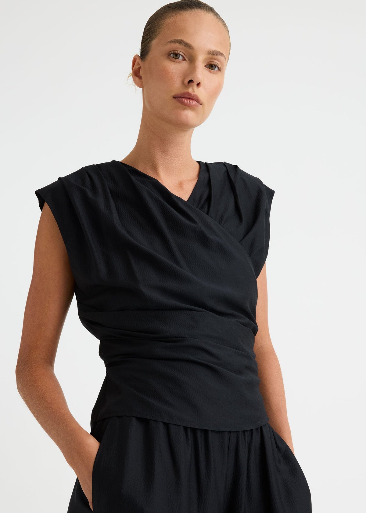 A woman wearing a black draped wrap-style top with cap sleeves and a matching black skirt, photographed against a clean light grey studio background.