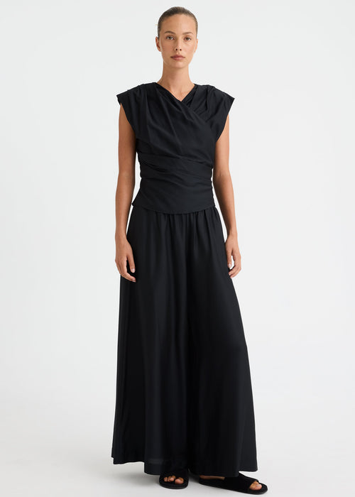 A model wearing a black sleeveless maxi dress with a draped, gathered neckline and a flowing skirt, standing against a plain light grey studio background.