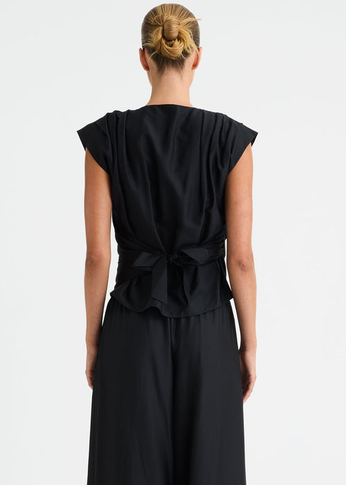 A woman viewed from the back wearing a black sleeveless gathered top, draped bodice and wide legs, standing against a plain light grey studio background.