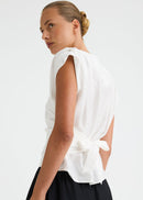 A woman shown from the back wearing a white sleeveless top with a tied waist detail and black wide-leg pants, photographed against a plain light grey studio background.