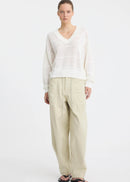 A woman stands against a plain white studio background wearing a white long-sleeve knit top with subtle horizontal texture details, beige wide-leg drawstring pants, and black sandals.