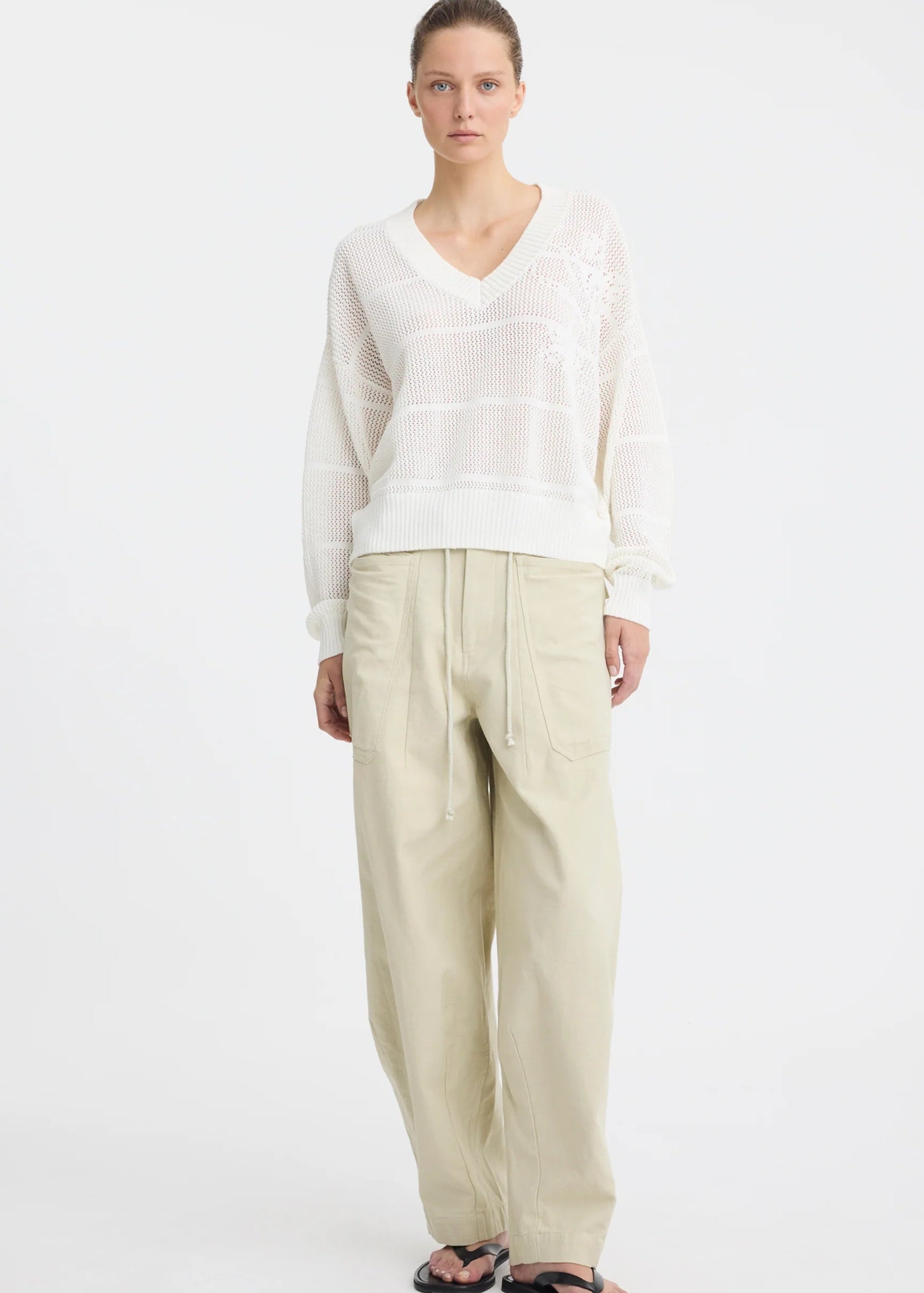 A woman stands against a plain white studio background wearing a white long-sleeve knit top with subtle horizontal texture details, beige wide-leg drawstring pants, and black sandals.