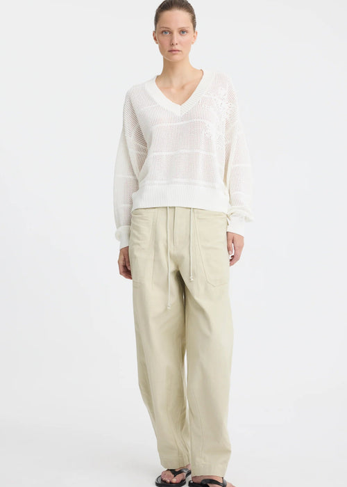 A woman stands against a plain white studio background wearing a white long-sleeve knit top with subtle horizontal texture details, beige wide-leg drawstring pants, and black sandals.