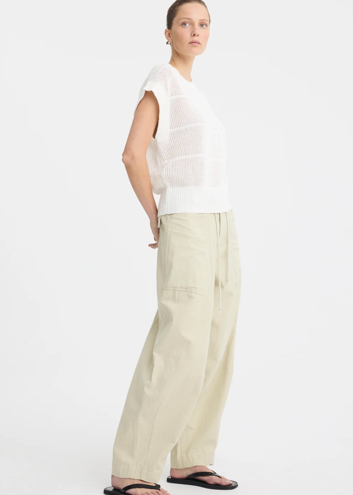 A woman stands against a plain white studio background wearing a white long-sleeve knit top with subtle horizontal texture details, beige wide-leg drawstring pants, and black sandals.