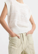 A woman stands against a plain white studio background wearing a white long-sleeve knit top with subtle horizontal texture details, beige wide-leg drawstring pants, and black sandals.