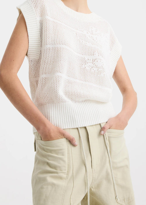 A woman stands against a plain white studio background wearing a white long-sleeve knit top with subtle horizontal texture details, beige wide-leg drawstring pants, and black sandals.
