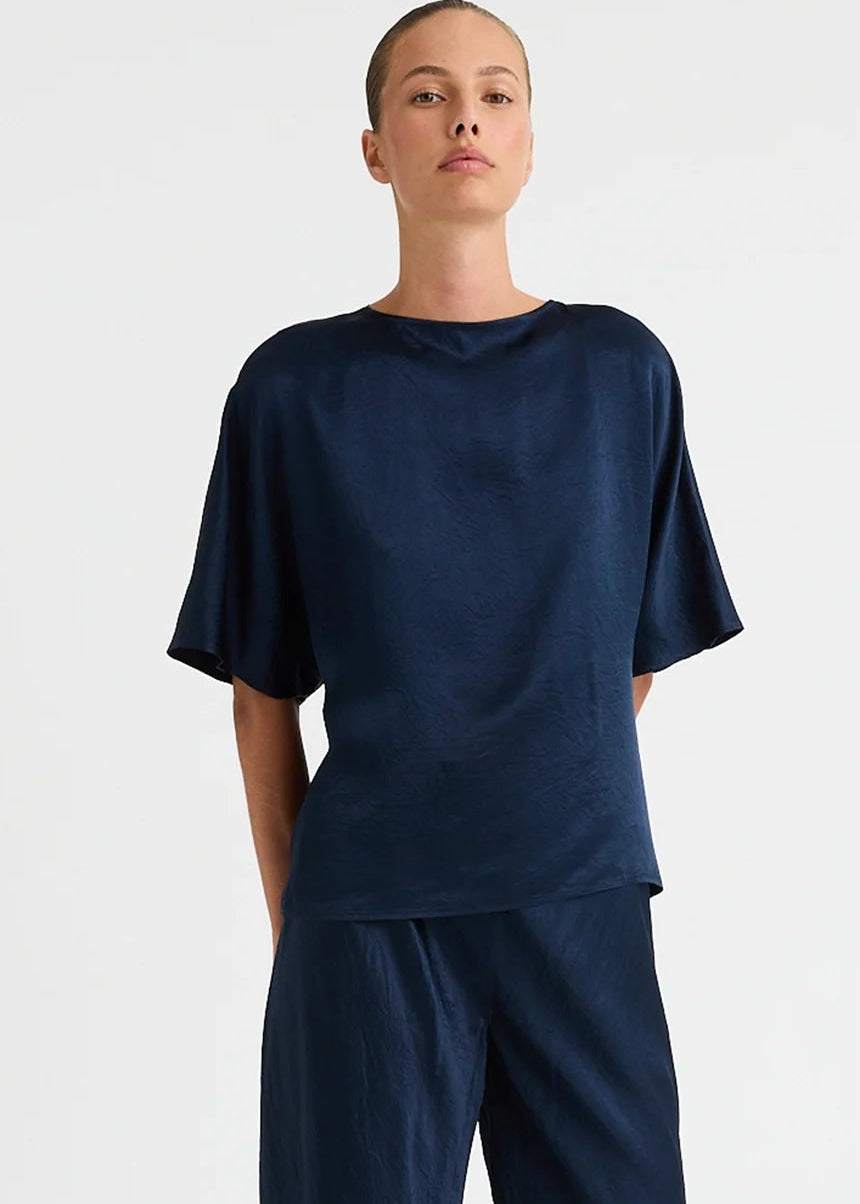 A model stands against a plain light grey studio background wearing a dark navy blue two-piece lounge set consisting of a short-sleeve top and matching wide-leg pants.