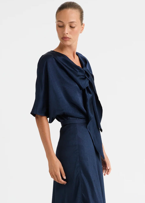 A model stands against a plain light grey studio background wearing a dark navy blue two-piece lounge set consisting of a short-sleeve top and matching wide-leg pants.