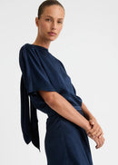 A model stands against a plain light grey studio background wearing a dark navy blue two-piece lounge set consisting of a short-sleeve top and matching wide-leg pants.