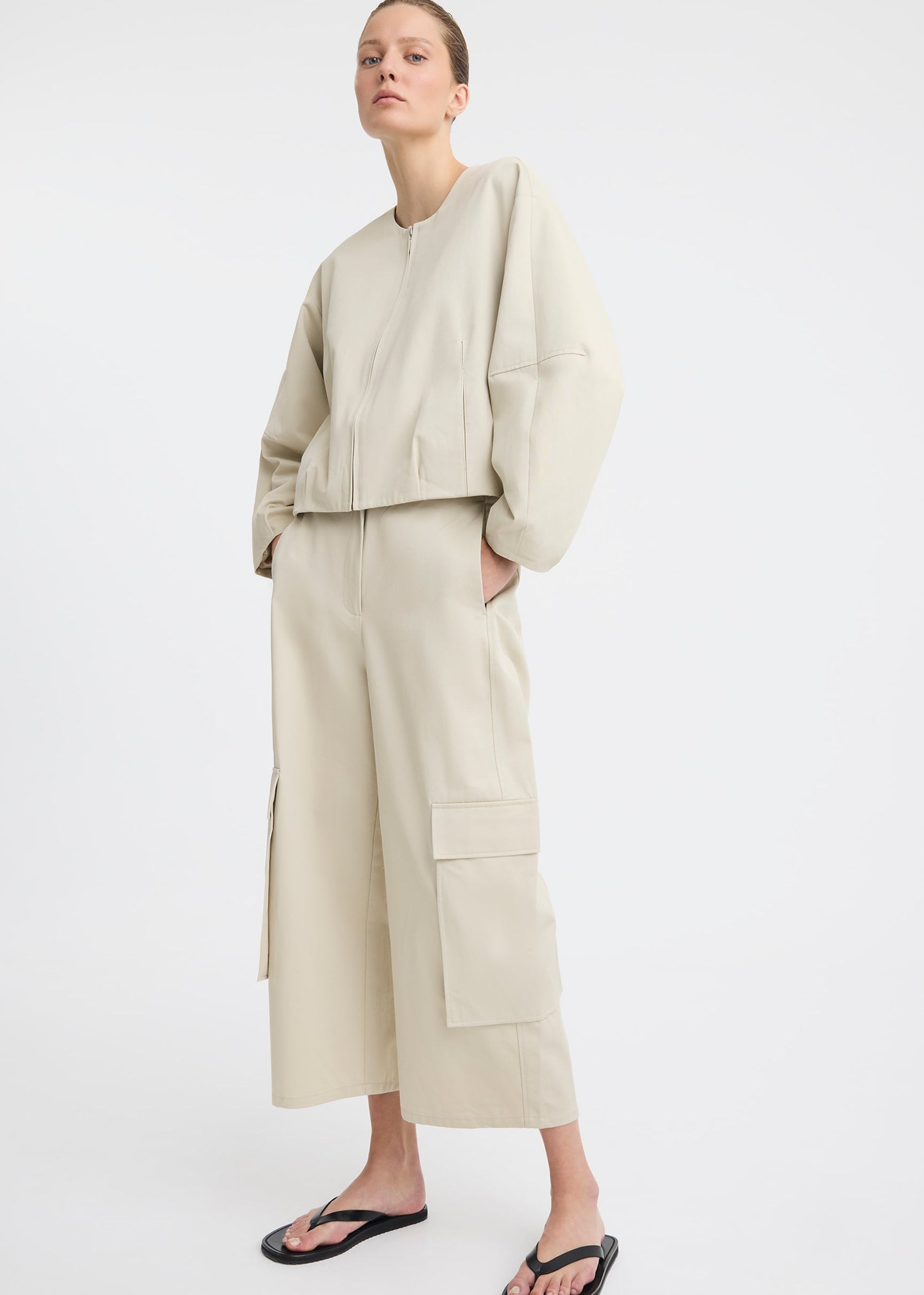 A model wearing a light beige two-piece outfit consisting of a cropped long-sleeve top and matching wide-leg cargo pants, paired with black flip-flop sandals, against a plain white studio background.