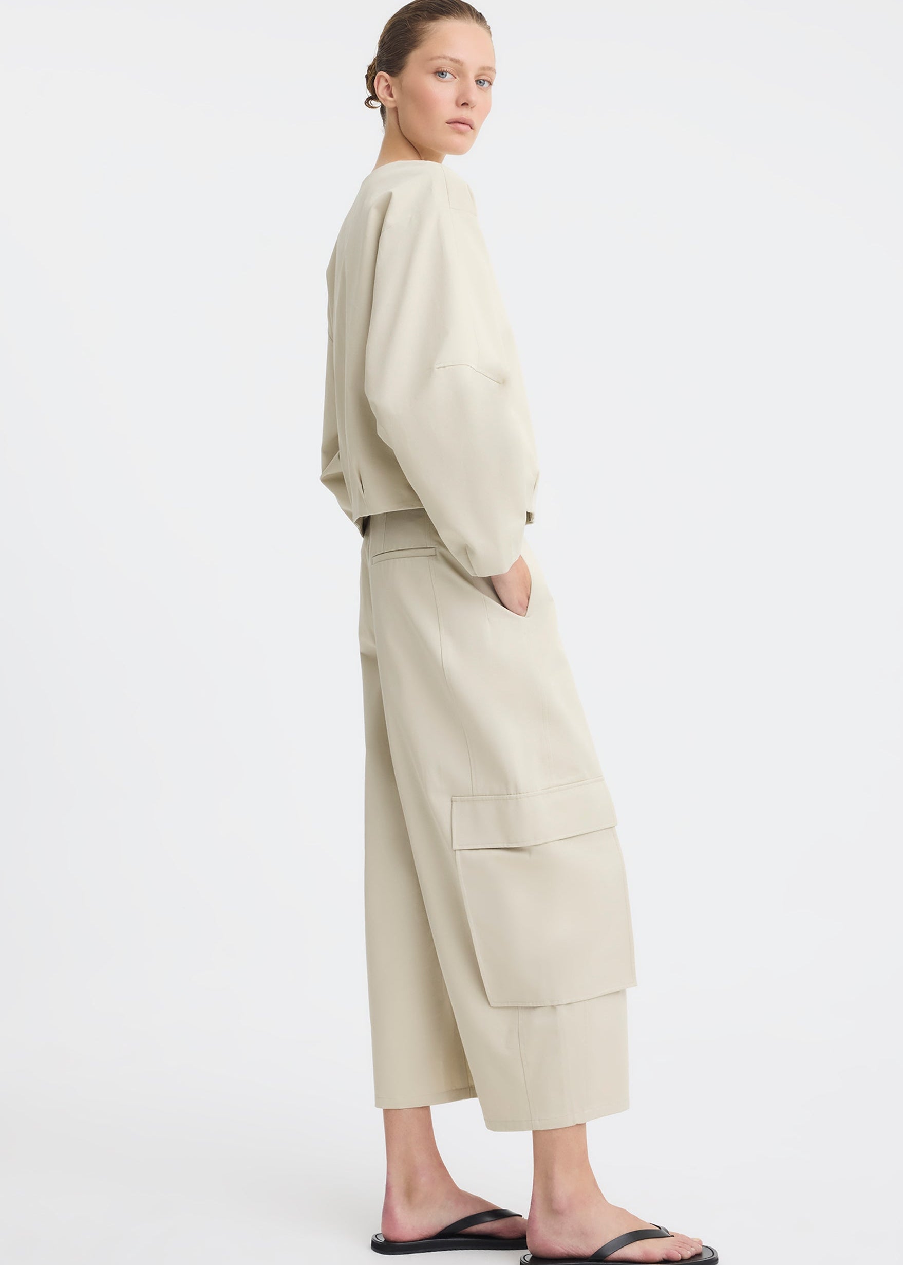 A model wearing a light beige two-piece outfit consisting of a cropped long-sleeve top and matching wide-leg cargo pants, paired with black flip-flop sandals, against a plain white studio background.