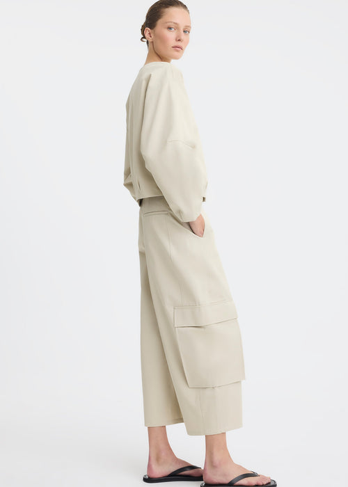 A model wearing a light beige two-piece outfit consisting of a cropped long-sleeve top and matching wide-leg cargo pants, paired with black flip-flop sandals, against a plain white studio background.