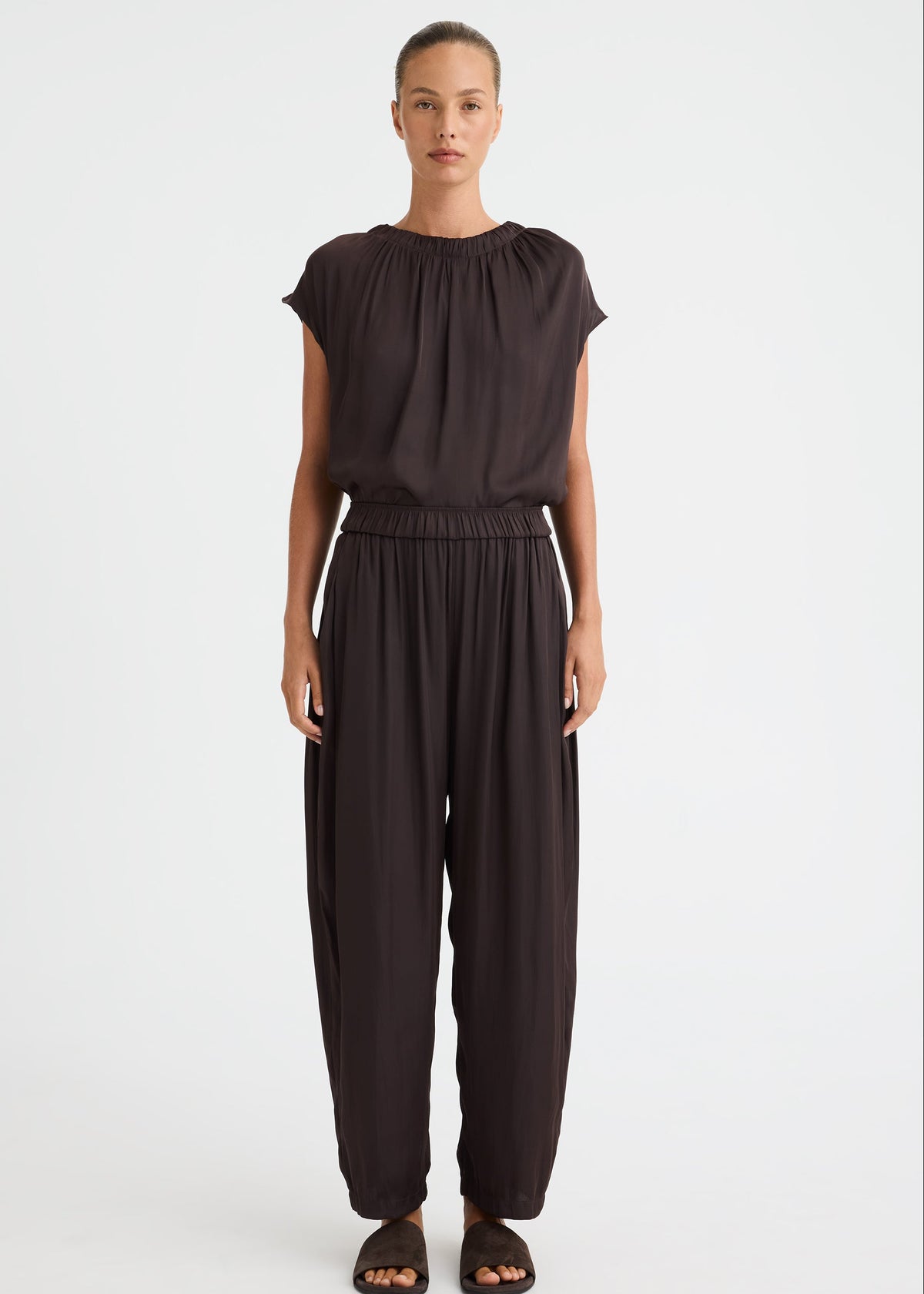 A model stands against a light grey studio background wearing a dark brown, loose-fitting two-piece outfit consisting of a short-sleeve top with a gathered neckline and matching wide-leg pants. The model is also wearing dark brown slide sandals.