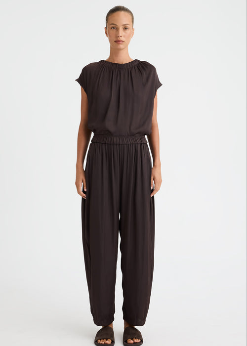 A model stands against a light grey studio background wearing a dark brown, loose-fitting two-piece outfit consisting of a short-sleeve top with a gathered neckline and matching wide-leg pants. The model is also wearing dark brown slide sandals.