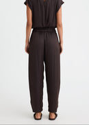 A woman viewed from the back wearing a dark brown short-sleeve jumpsuit with a small keyhole cut out at the back of the neckline and wide-leg pants, standing against a plain white studio background.