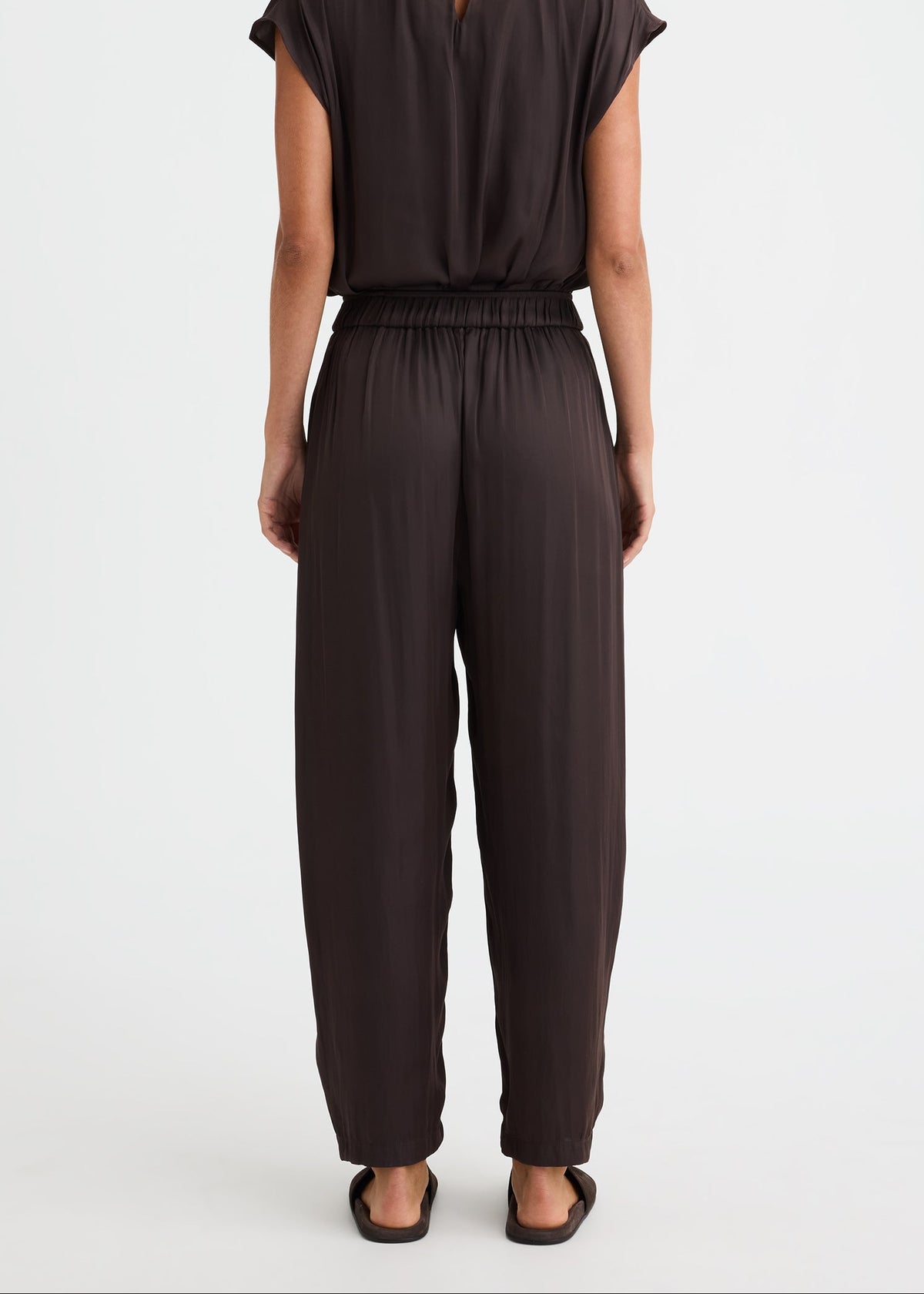 A woman viewed from the back wearing a dark brown short-sleeve jumpsuit with a small keyhole cut out at the back of the neckline and wide-leg pants, standing against a plain white studio background.