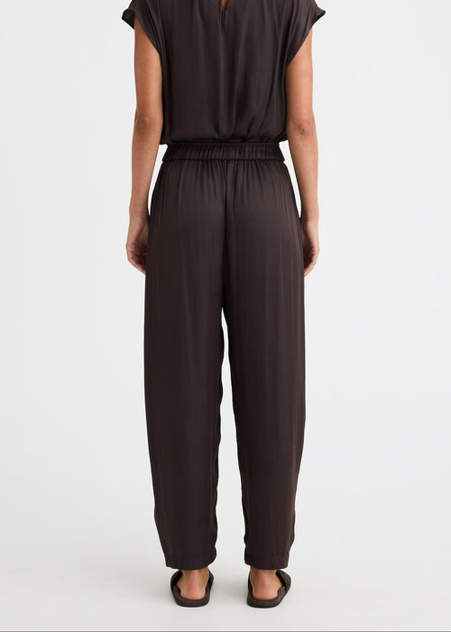 A woman viewed from the back wearing a dark brown short-sleeve jumpsuit with a small keyhole cut out at the back of the neckline and wide-leg pants, standing against a plain white studio background.