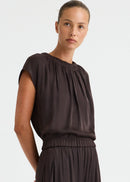 A woman modelling a dark brown two-piece outfit consisting of a short-sleeve top with a gathered neckline and matching loose-fitting wide-leg pants, paired with dark brown slide sandals against a plain white studio background.