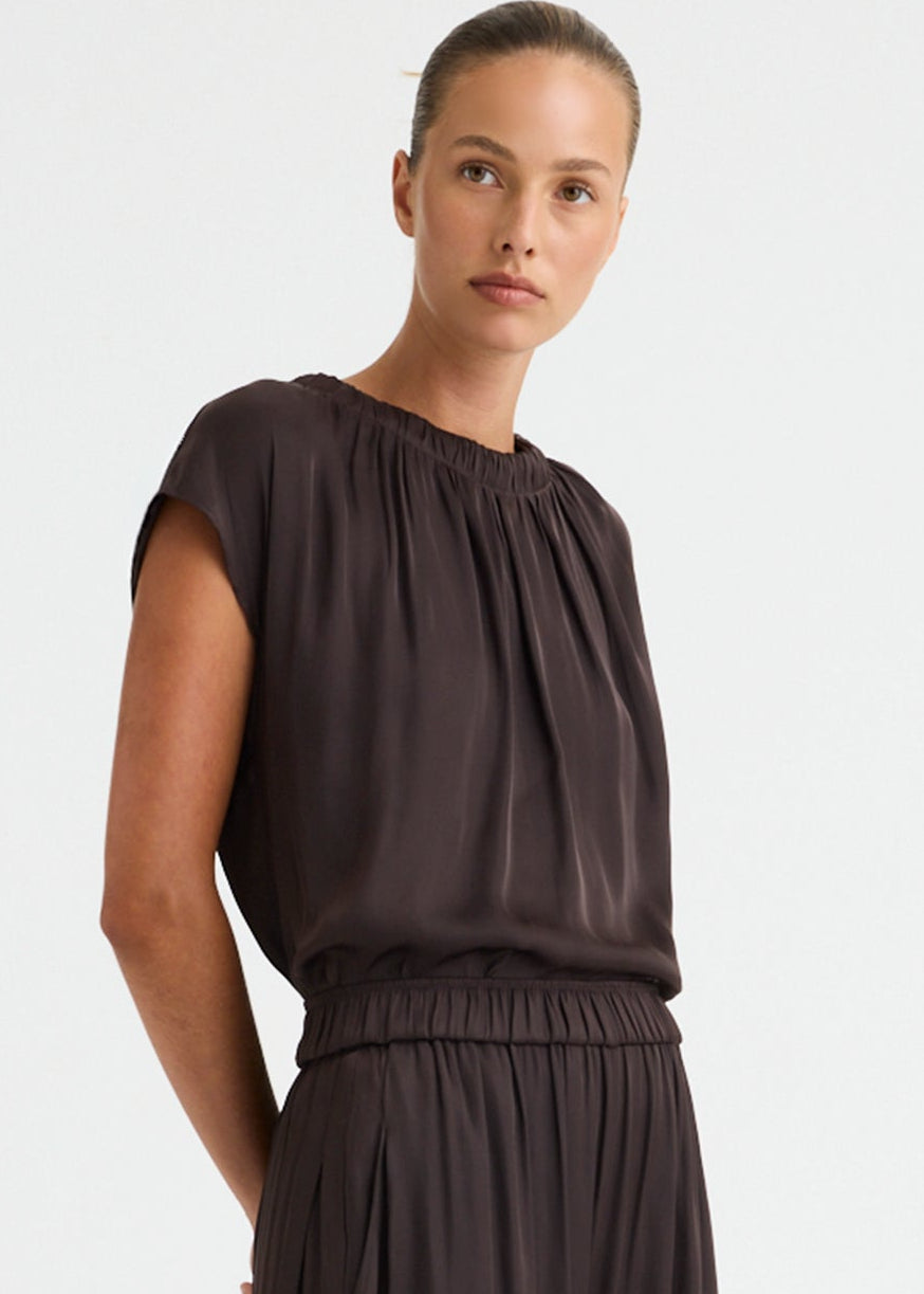 A woman modelling a dark brown two-piece outfit consisting of a short-sleeve top with a gathered neckline and matching loose-fitting wide-leg pants, paired with dark brown slide sandals against a plain white studio background.
