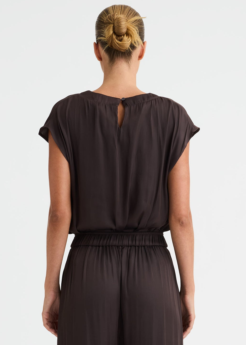 A woman viewed from the back wearing a dark brown short-sleeve jumpsuit with a small keyhole cut out at the back of the neckline and wide-leg pants, standing against a plain white studio background.