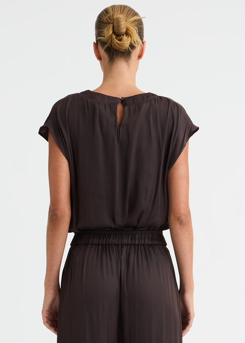 A woman viewed from the back wearing a dark brown short-sleeve jumpsuit with a small keyhole cut out at the back of the neckline and wide-leg pants, standing against a plain white studio background.
