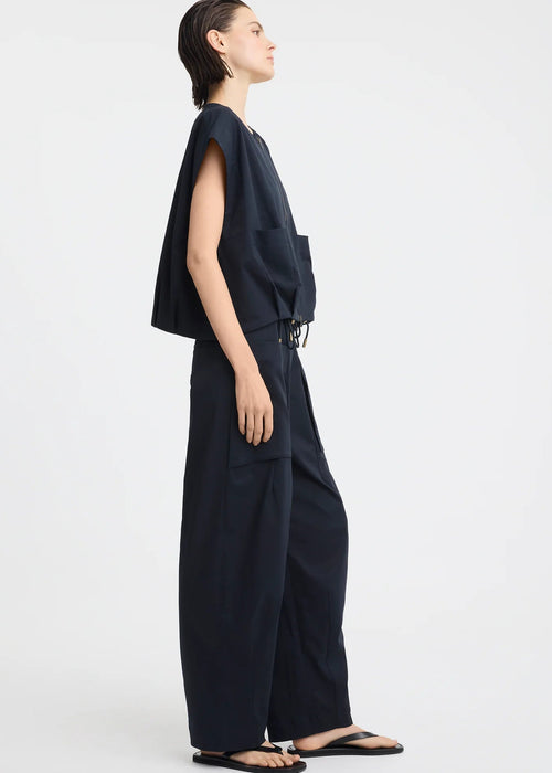 A model stands against a light grey studio background wearing a dark navy sleeveless top with a front zipper and matching dark navy wide-leg pants with large front pockets.