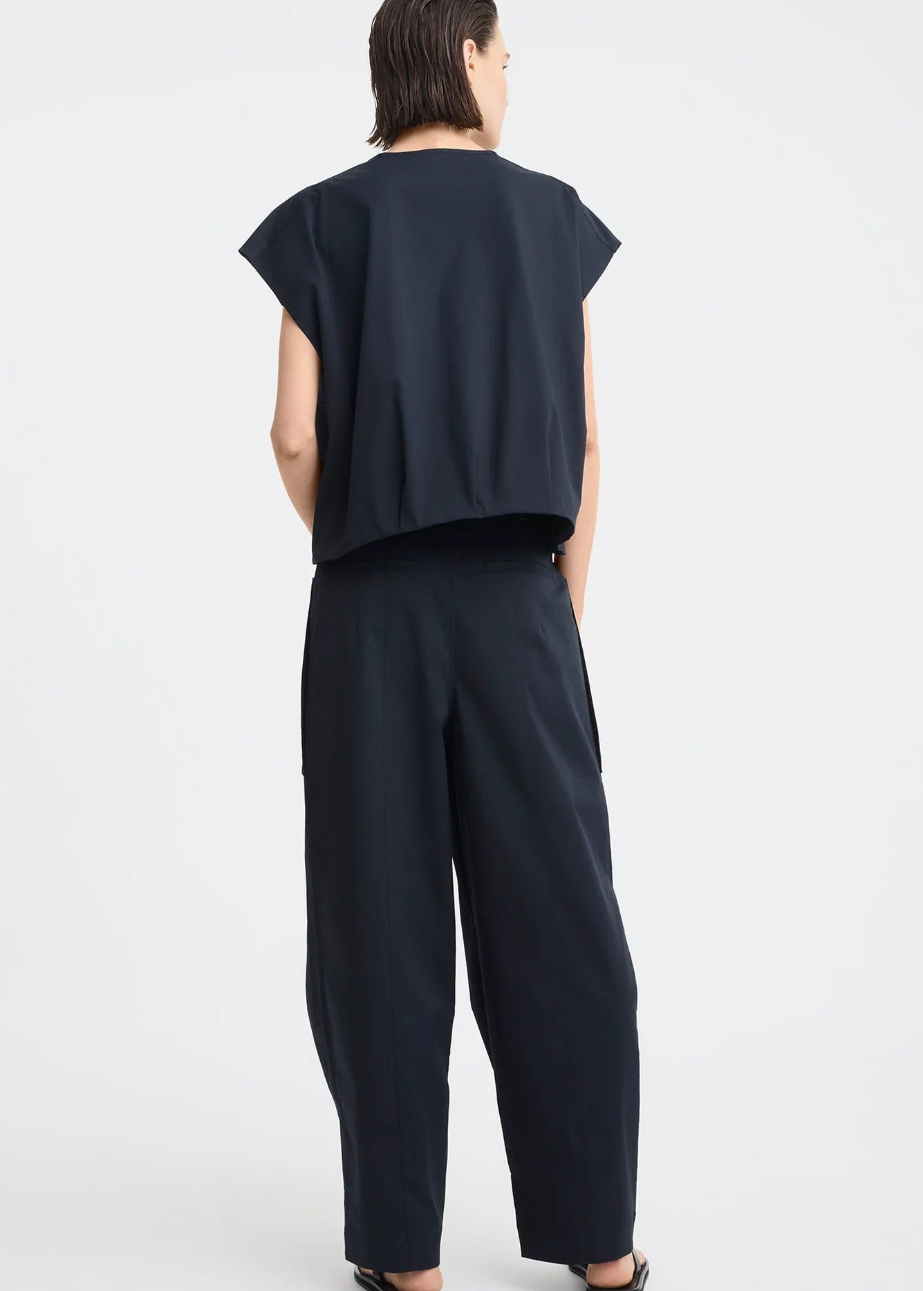 A model stands against a light grey studio background wearing a dark navy sleeveless top with a front zipper and matching dark navy wide-leg pants with large front pockets.