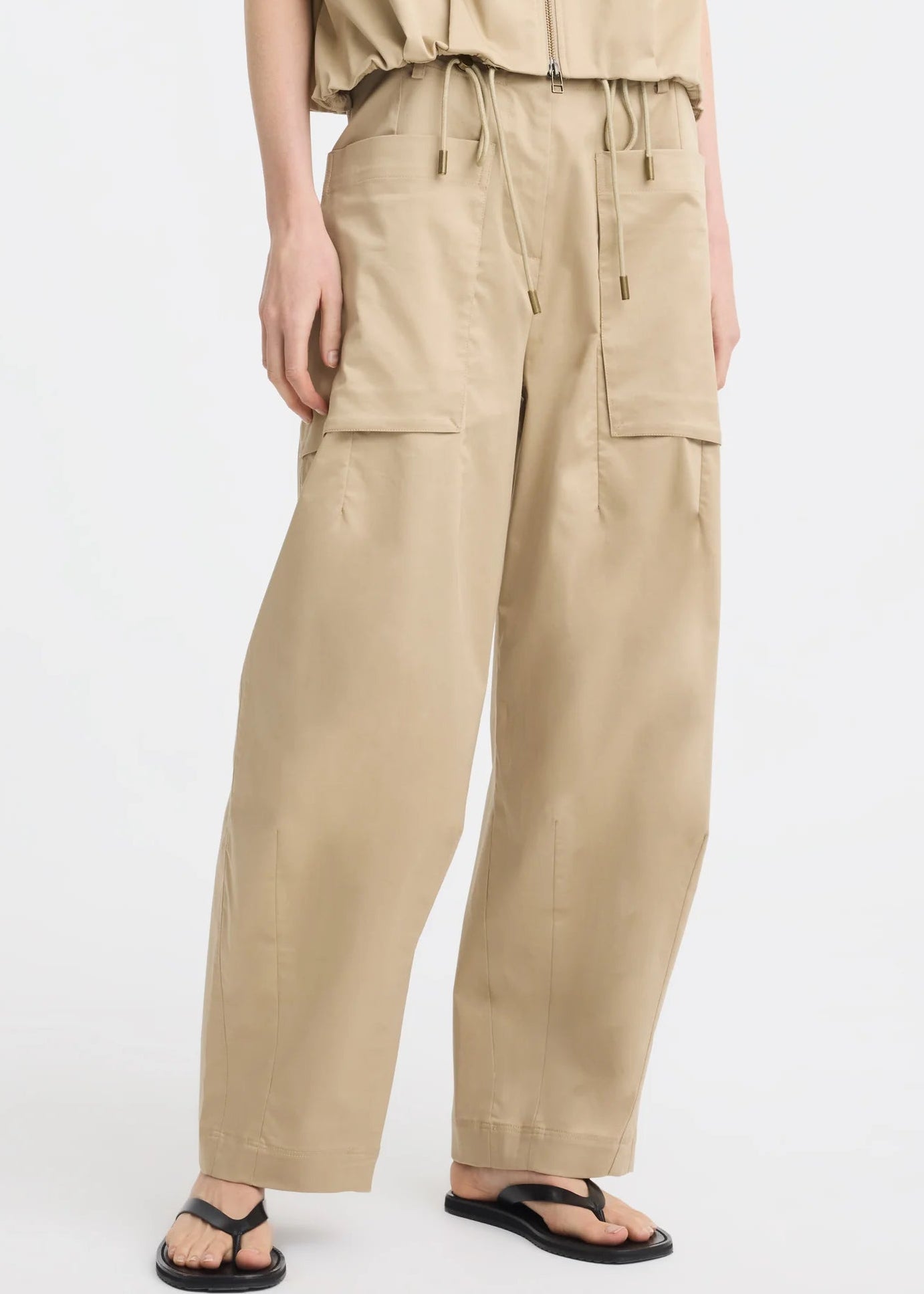 A model wearing a beige sleeveless top and matching wide-leg cargo pants, paired with black flip-flop sandals, photographed against a plain white studio background.
